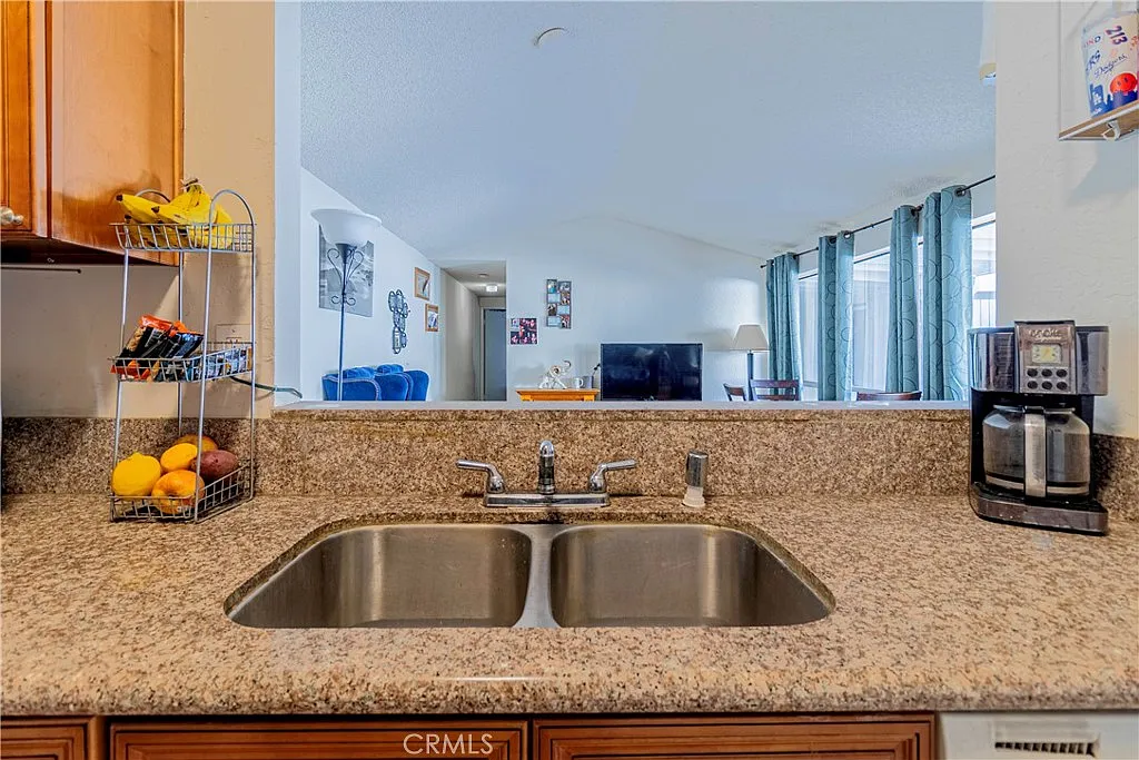 Kitchen sink with granite countertop, fruit basket, and living area with blue couches and large windows in the background.