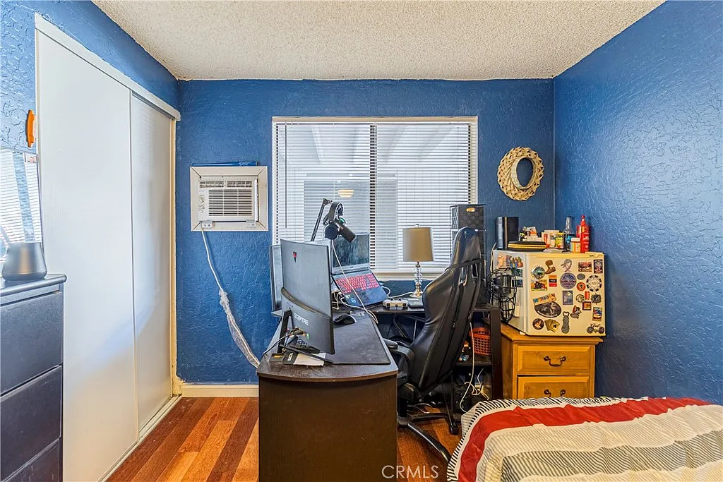 Small room with blue walls, desk with computer, chair, window air conditioner, mirror, and dresser with items.