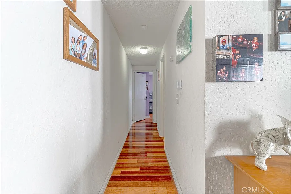 Narrow hallway with wooden floor, white walls, framed photos on left, and decorative items on table to right.