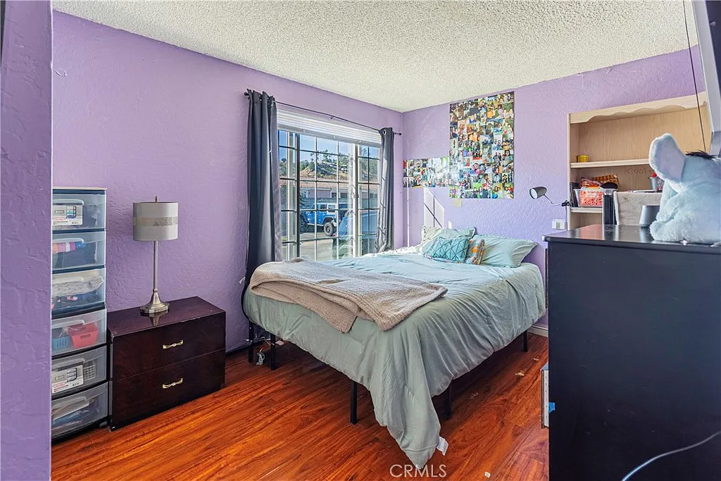 Bedroom with purple walls, bed with light blue bedding, nightstand, window view, photo collages, and storage bins beside bed.