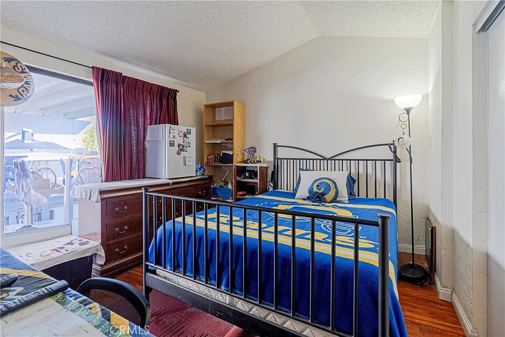Bedroom with bed with blue and yellow bedding, wooden dresser, desk with items, small refrigerator, and window with red curtains.