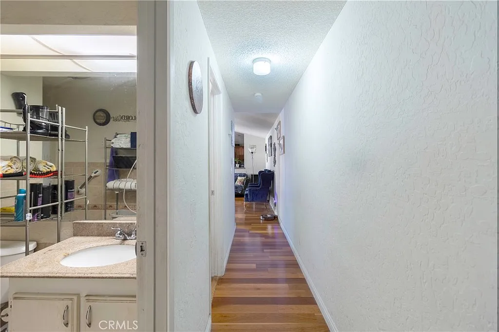 Hallway with light walls, wooden floor, doors to rooms, shelves with items, and framed pictures at the end.