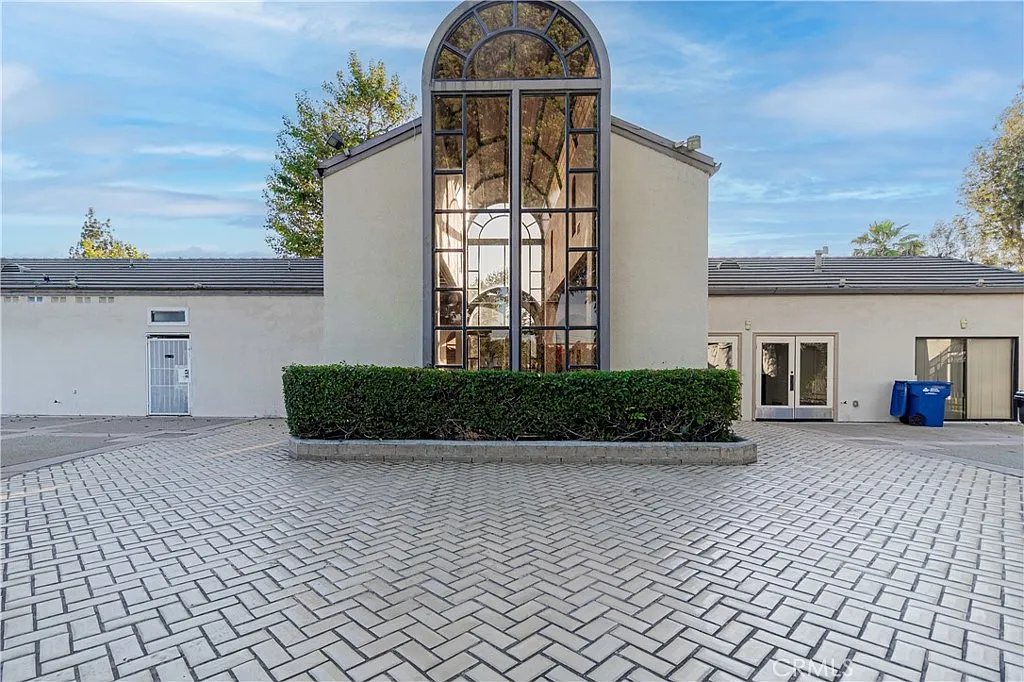 Modern building with large arched window, greenery, and brick-paved area.