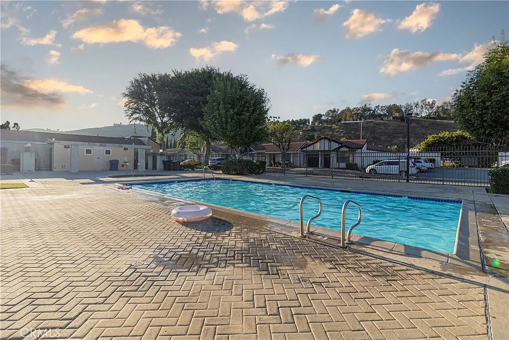 Swimming pool with light blue water, tan brick patio, trees, nearby houses, and floating inflatable ring.