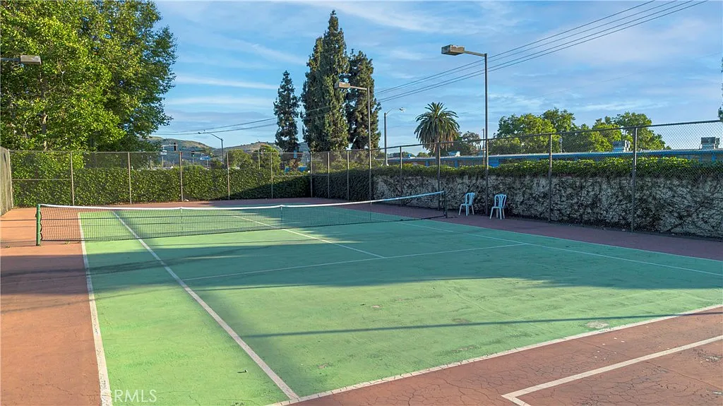 Empty tennis court with green and red surface, surrounded by trees and fence, with chairs nearby.