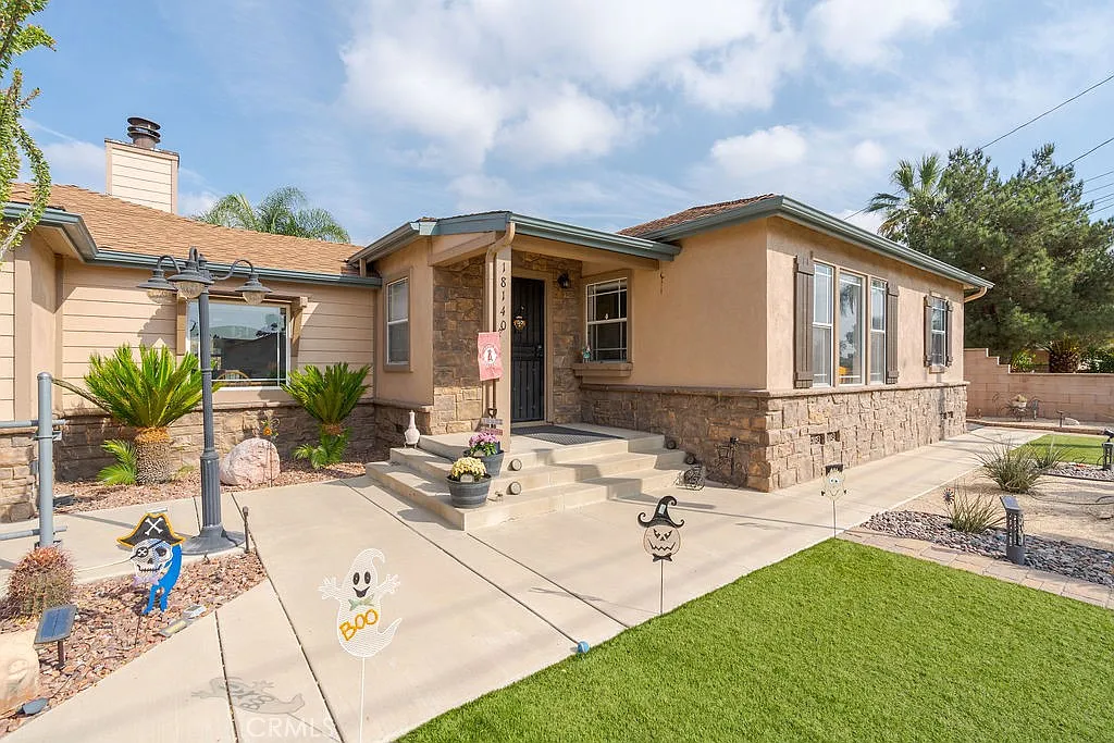 single-story home with a stone facade, front porch, Halloween yard decorations, green lawn, palm trees, and shrubs.
