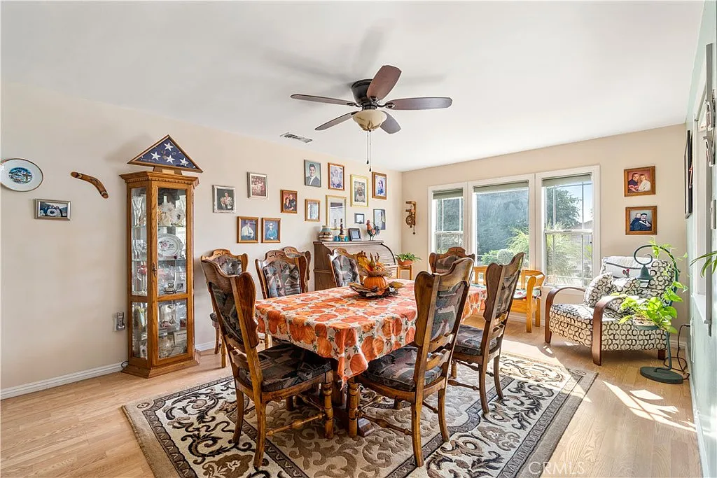 Dining room with a large wooden table, patterned tablecloth, ornate chairs, glass display cabinet, family photos on the walls, and a cozy armchair near the windows.