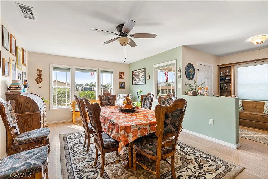 Dining area with a large table, wooden chairs, ceiling fan, windows, framed pictures on the walls, and a patterned tablecloth.