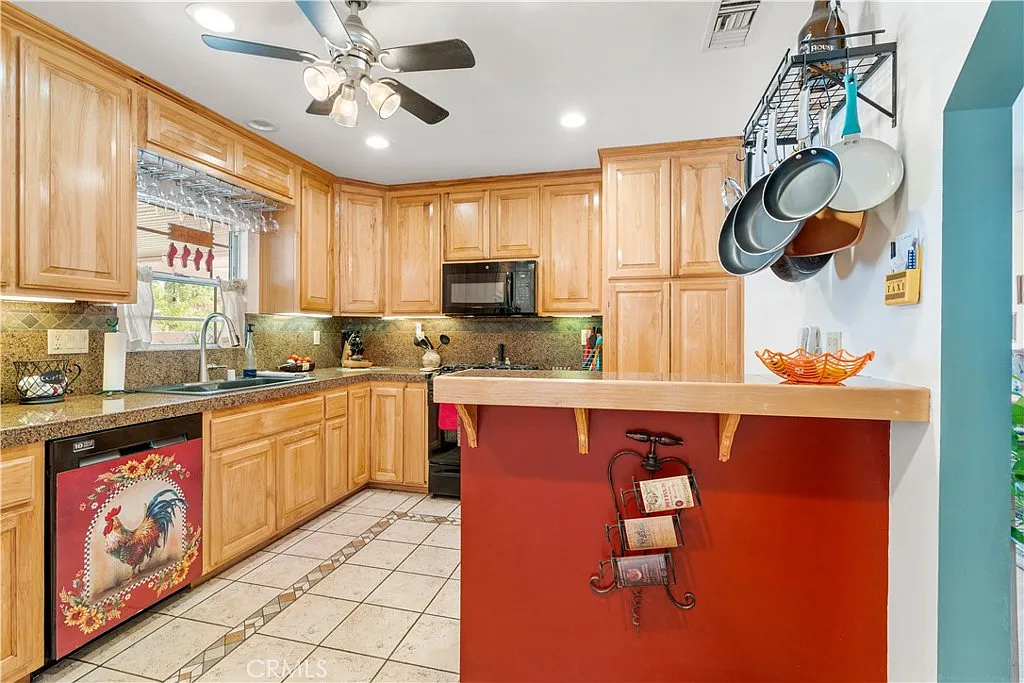 A kitchen with wooden cabinets, a countertop with a red wall, kitchen appliances, a rooster decoration on the dishwasher, and hanging pots above the counter.