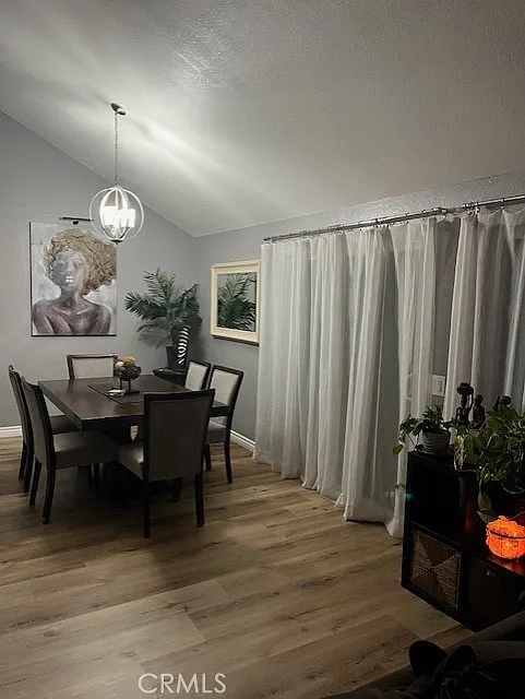 Dining room with a wooden table, six chairs, wall artwork, sheer white curtains, corner plants, pendant lamp, and light wood floors.