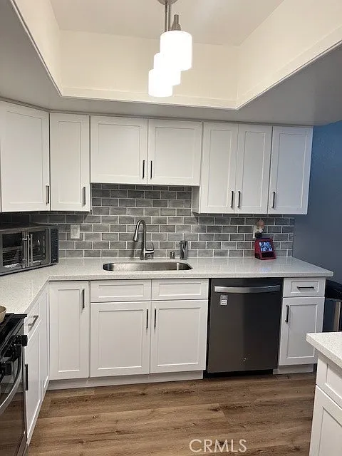 Kitchen with white cabinets, stainless steel sink, gray tile backsplash, dishwasher, microwave, and wooden floors.