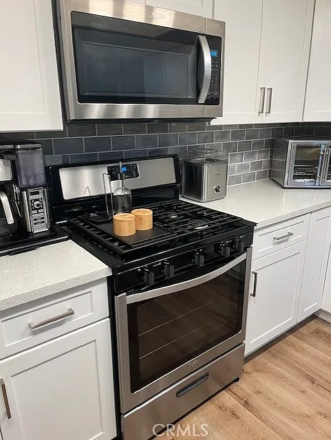 Kitchen with stainless steel gas stove and oven, microwave above, white cabinets, gray tile backsplash, and appliances on the counter.