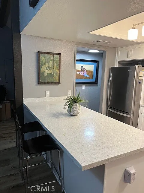 Kitchen with white countertop, bar seating, wall art, TV in the background, stainless steel appliances, and light-colored cabinets.
