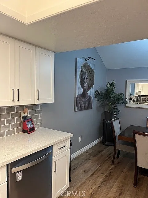 Kitchen with white cabinets, stainless steel dishwasher, gray wall with framed artwork, and a dining area with table and chairs in the background.