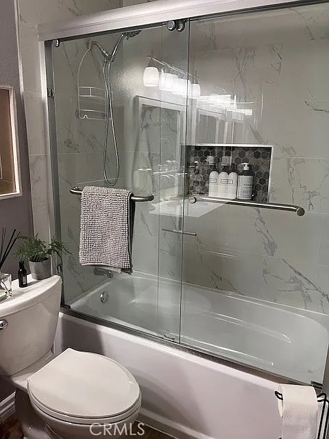Bathroom with glass shower, bathtub, toilet, shelf bottles, and towel bar.