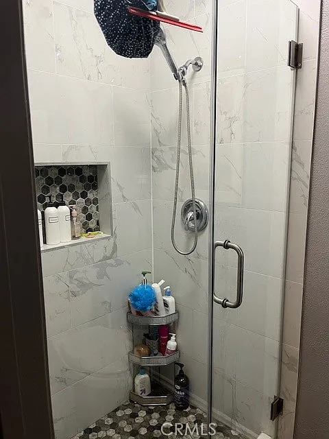 A modern shower with glass doors, rainfall showerhead, marble tiles, and a small shelf with toiletries.