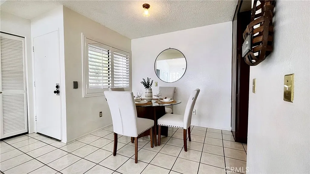 Small dining area with round table, white chairs, circular wall mirror, light walls, and window shutters.