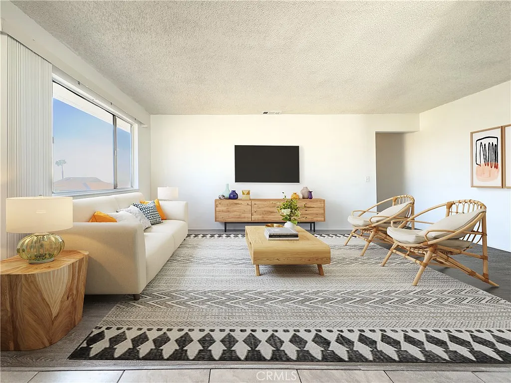 Living room with white sofa, colorful pillows, wooden coffee table, wicker chairs, TV on media console, large windows, and patterned rug.