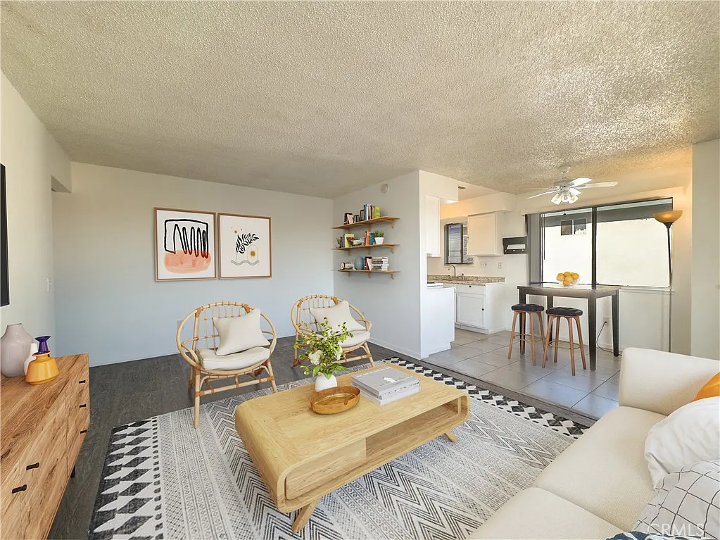Living room with coffee table on patterned rug, two rattan chairs, wall artwork, and kitchen in the background.