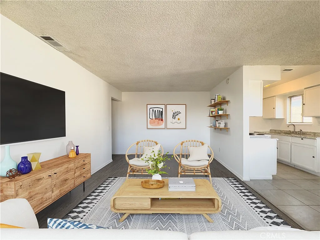 Living room with beige sofa, wooden media console, rattan chairs, coffee table, large tv, plants, framed artwork, and open kitchen in the background.
