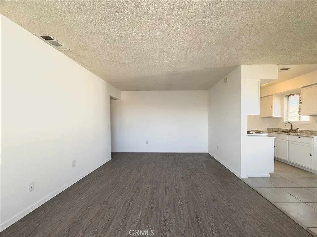 Empty living room with light walls, wooden floor, and open kitchen in the background.
