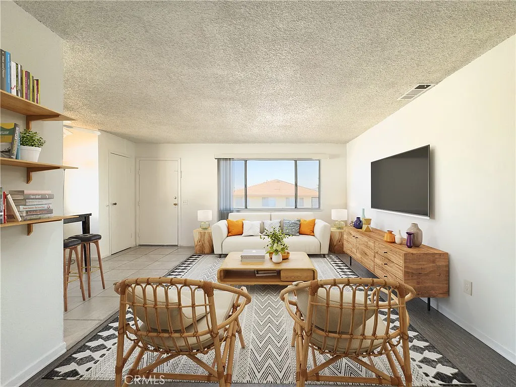 Living room with white sofa, orange and yellow pillows, rattan chairs, wooden coffee table, media console, large tv, and windows with natural light.