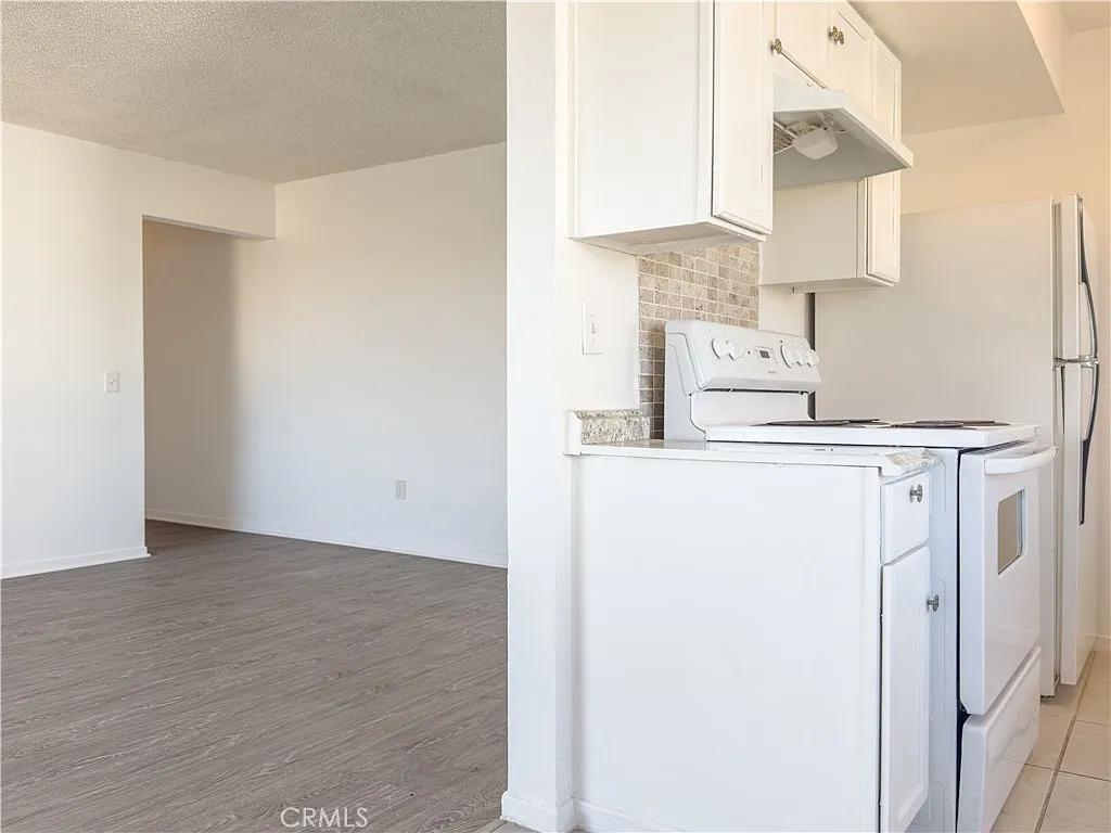 Empty kitchen with white cabinets, stove, refrigerator, and open adjacent room with light-colored floor.