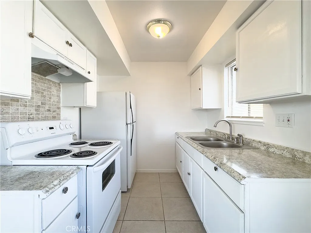 Kitchen with white cabinets, stainless steel sink, stove, oven, refrigerator, and granite countertops by a window.