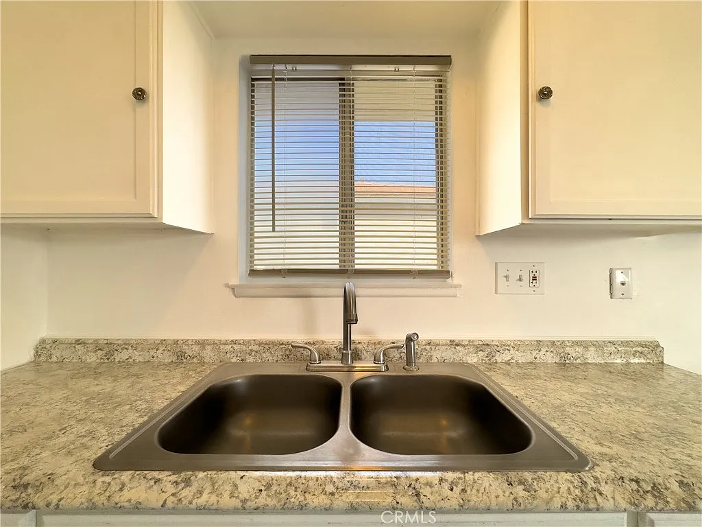 Kitchen sink with double basin and granite countertop under a window with blinds.