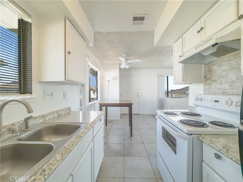 Kitchen with white cabinets, countertop, double sink, stove, small table, windows, and tile floor.