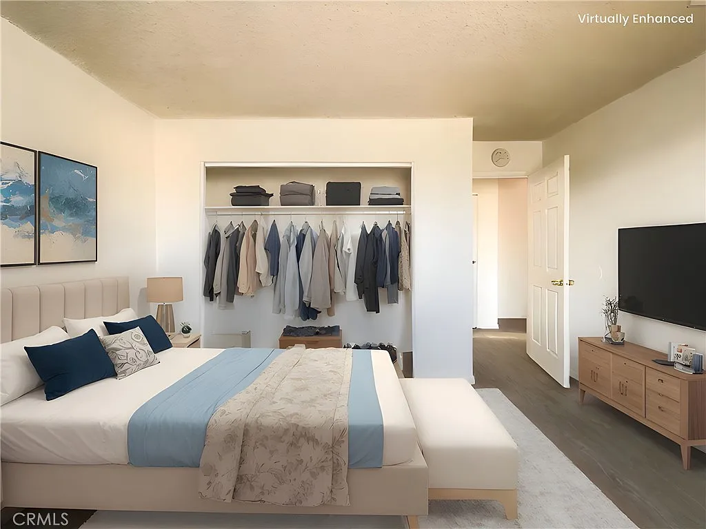 bedroom with light blue bed, beige headboard, bench, closet with clothes, TV stand, white walls, and natural light.