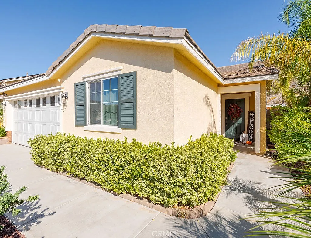 Single-story house real estate with tan walls, green shutters, porch, plants, and palm trees.