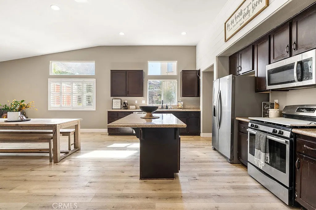 Kitchen with dark wooden cabinets, stainless steel appliances, large central island, dining table, and large windows