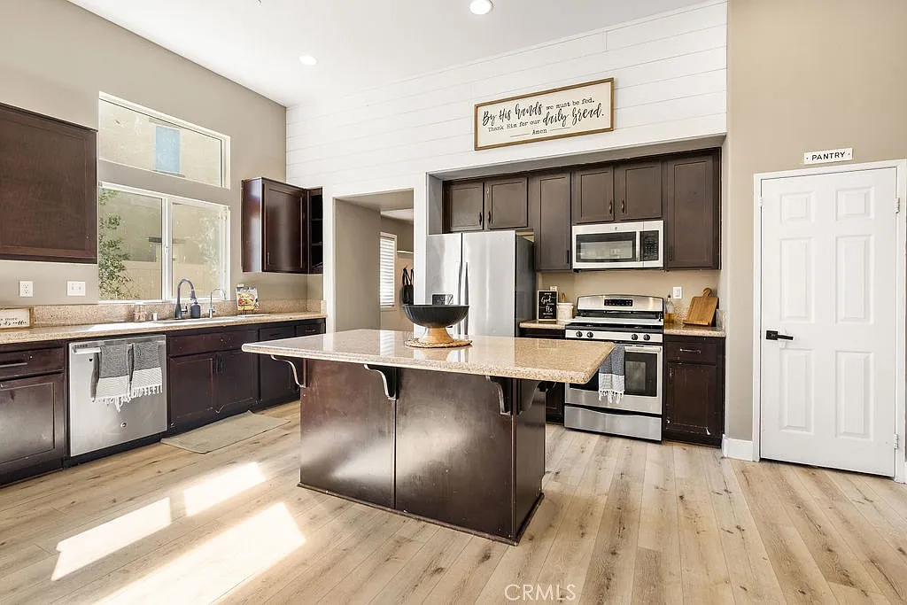 Kitchen with dark wooden cabinets, stainless steel appliances, central island with bowl, natural light from windows, decorative wall sign, and light wood floors.