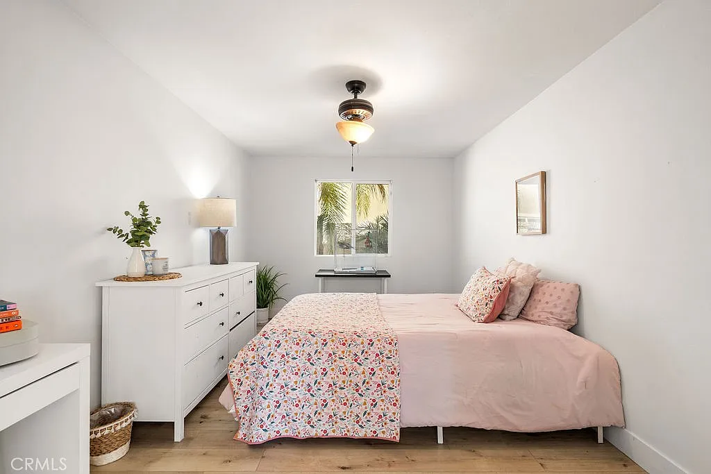 Bedroom with bed covered in pink floral blanket, white dresser, and window showing plants outside