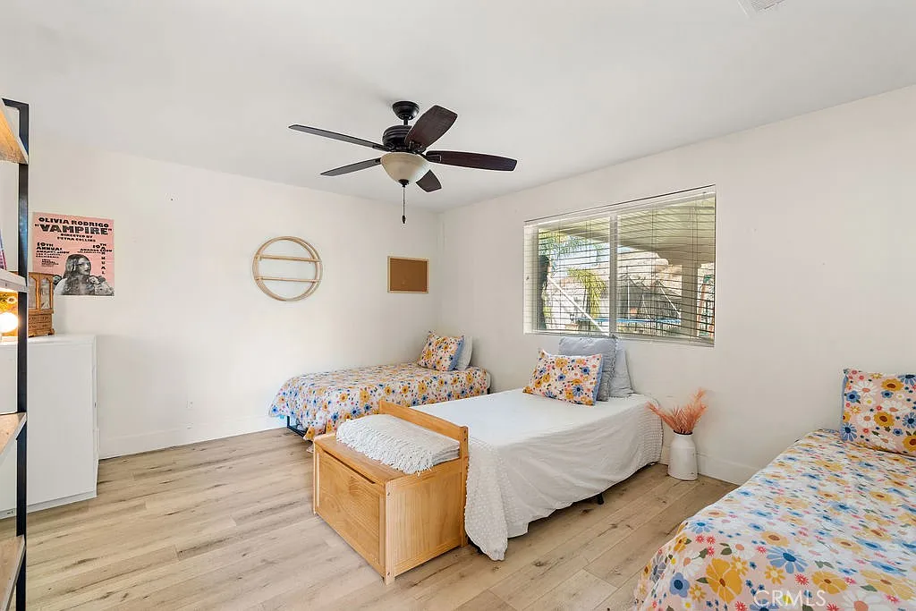 Room with three beds, ceiling fan, large windows, floral-patterned bedding, round mirror, and wooden storage unit.