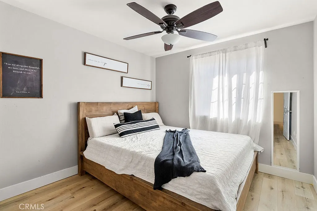 bedroom with wooden bed, white bedding, black and white throw, ceiling fan, large window with sheer curtains, and full-length mirror.