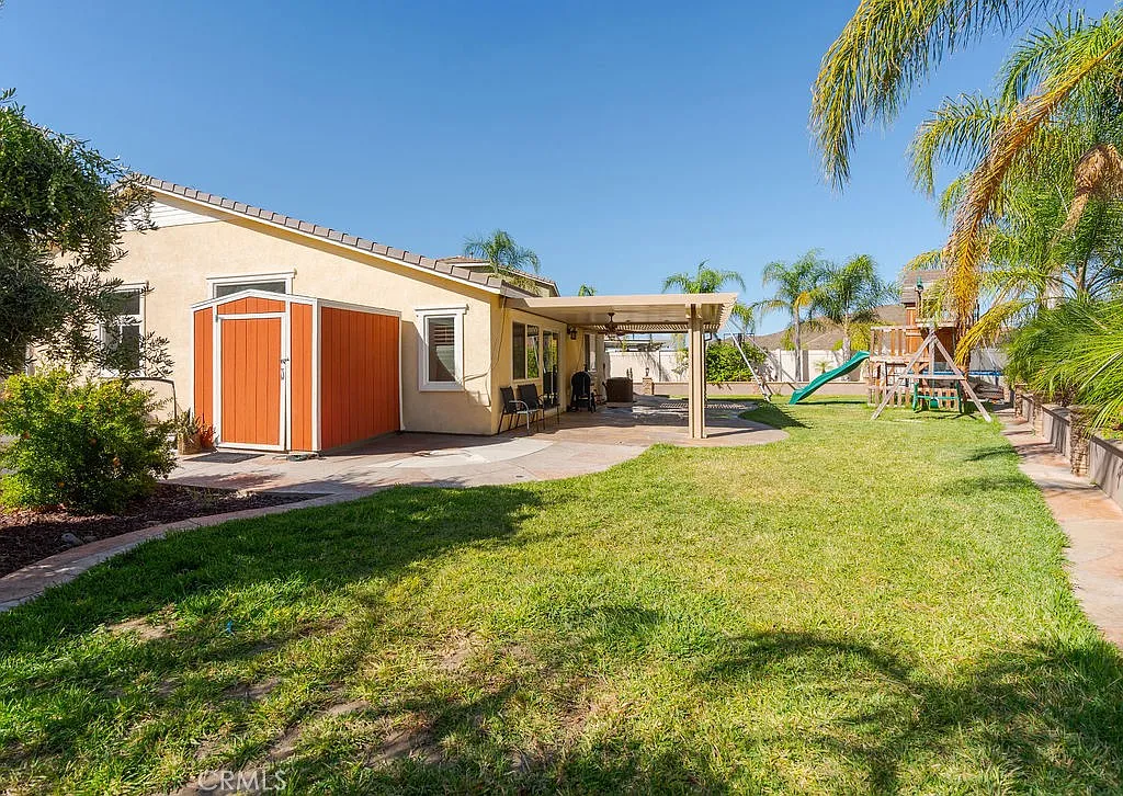 A backyard with a well-maintained lawn, patio with outdoor seating, playground, palm trees, and a light-colored house with an orange door.