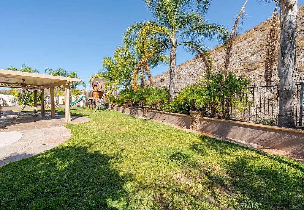 A landscaped backyard with green grass, palm trees, play structure, and a hillside.