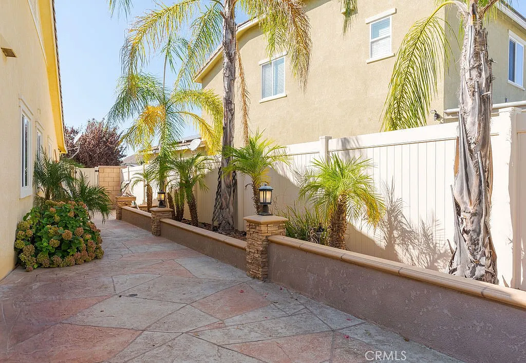 A narrow pathway lined with tropical plants and palm trees, light-colored fence, houses on both sides