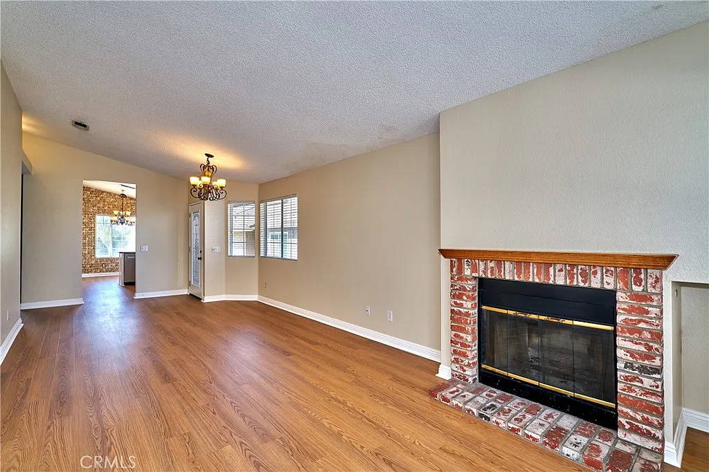 Living room with brick fireplace, hardwood floor, light walls, windows, chandelier, and doorway to another room.