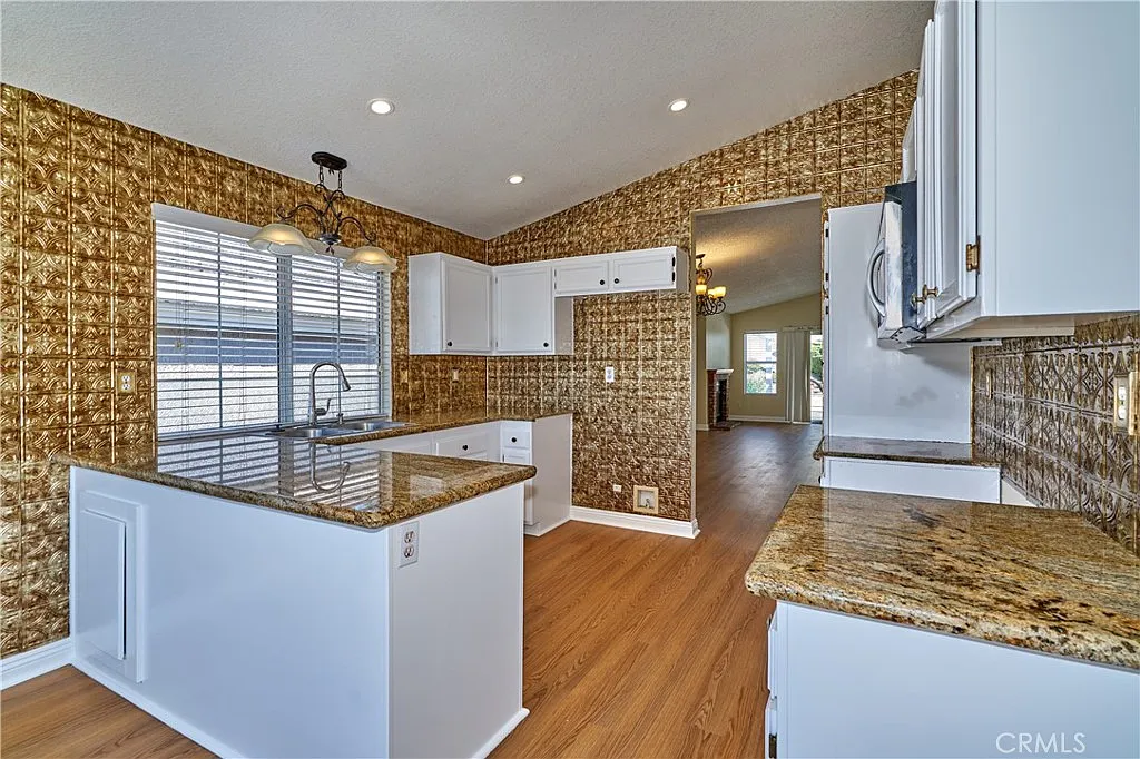 Kitchen with golden walls, white cabinets, granite countertop, wooden floor, window with blinds, and open doorway to living area.