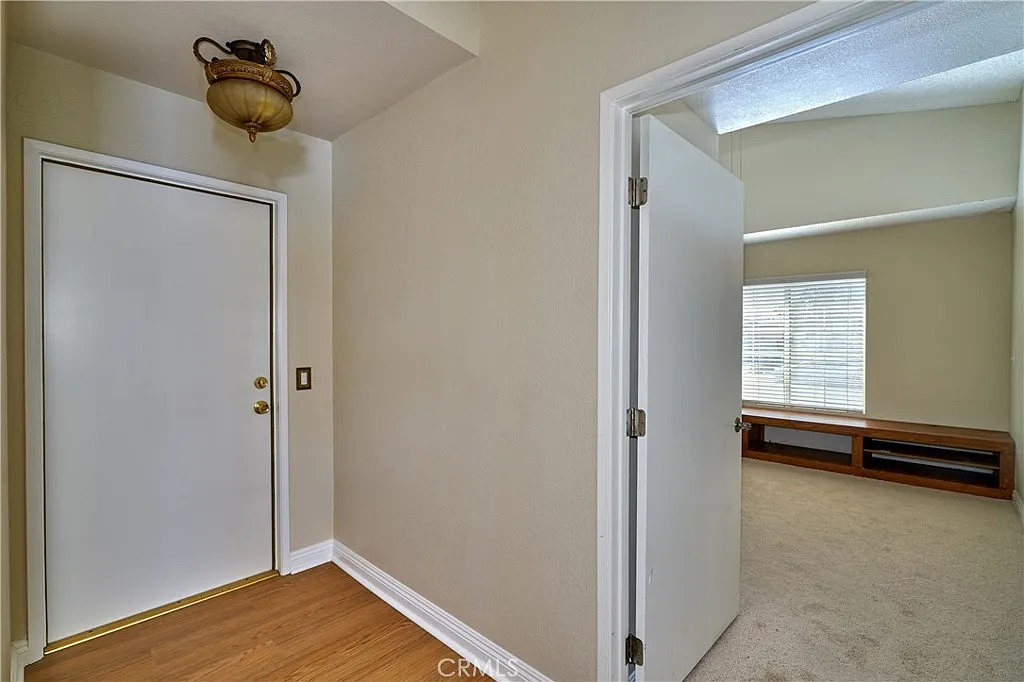 Hallway with white door, ceiling light, beige walls, and adjacent room with windows and sparse furniture.