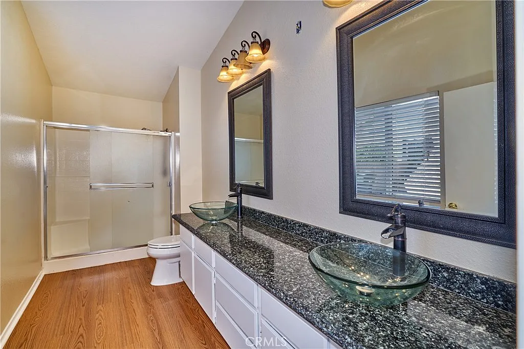 Bathroom with dual glass sinks, mirrors, walk-in shower, wooden floor, and beige walls.