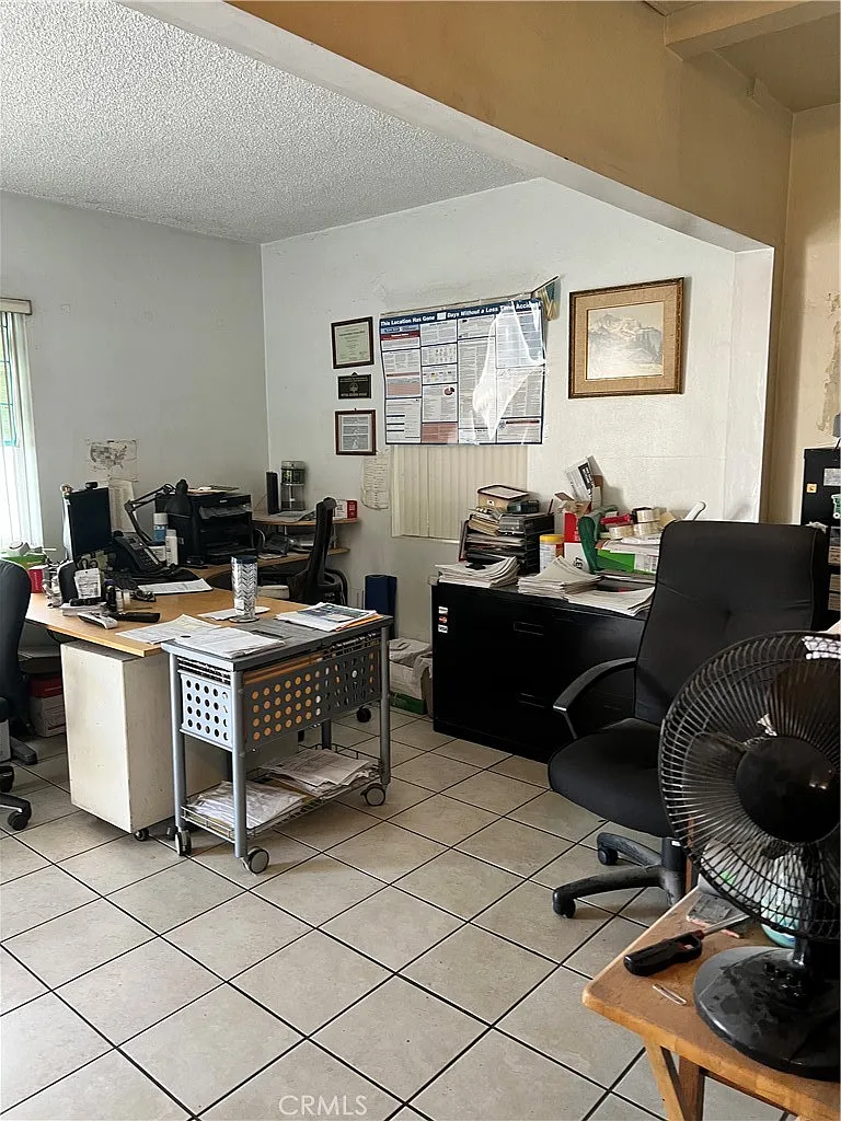 Office space with two desks, computer, paperwork, black filing cabinet, fan, and office supplies.