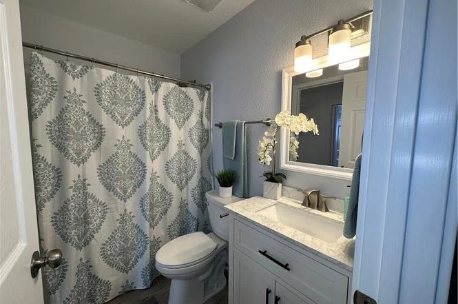Bathroom with shower and patterned curtain, toilet, vanity with sink and mirror, plants, and towels
