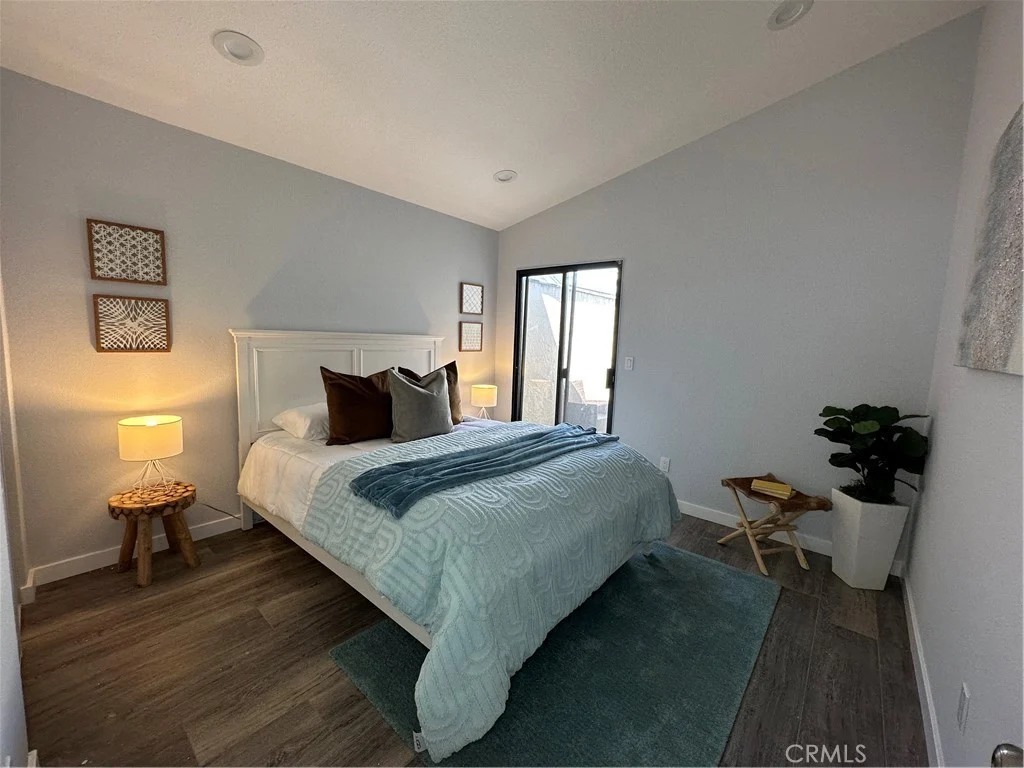 bedroom with white bed, decorative pillows, blue throw, bedside lamps, sliding glass door to outdoor space, light gray walls, wooden floor, plant, and small stool