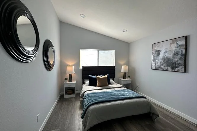 bedroom with queen bed, gray and blue bedding, two lamps on white nightstands, round wall mirrors, window with natural light, and abstract artwork above bed