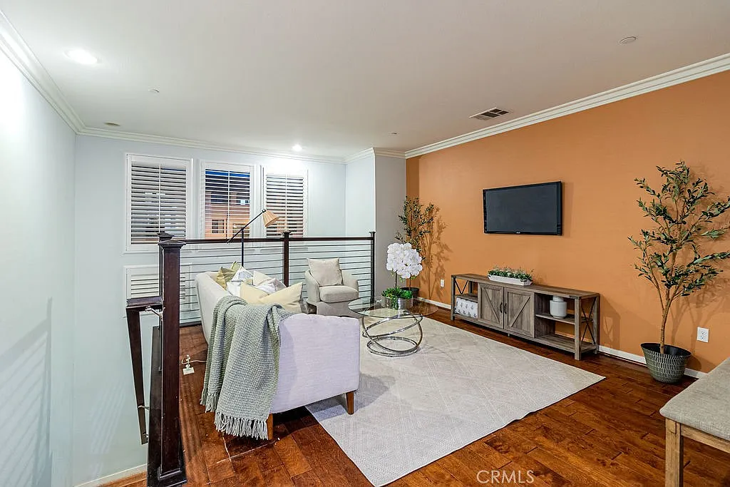 living room with gray sofa, glass coffee table, wall TV, plants, window shutters, and wood floor.