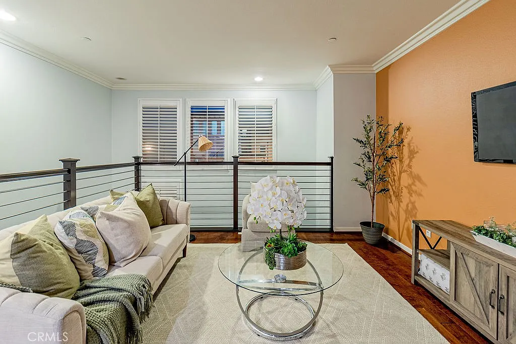 Living room with light sofa, pillows, round glass coffee table with white flowers, wooden tv stand, orange wall, large windows, and floor lamp.